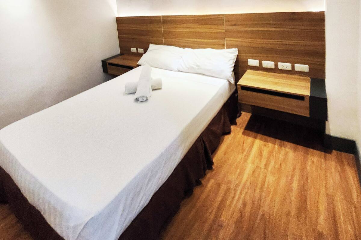 Una cama individual con cabecera de madera, ropa de cama blanca y una mesita de noche de madera con lámpara.
