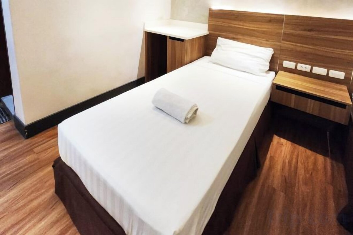 A neatly made bed with a white sheet and a rolled towel, a wooden nightstand with a lamp, and a wooden headboard.