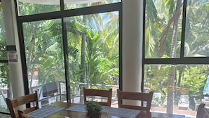 Comedor con mesa y sillas de madera, ventanas amplias con vista a palmeras y un ventilador de techo.