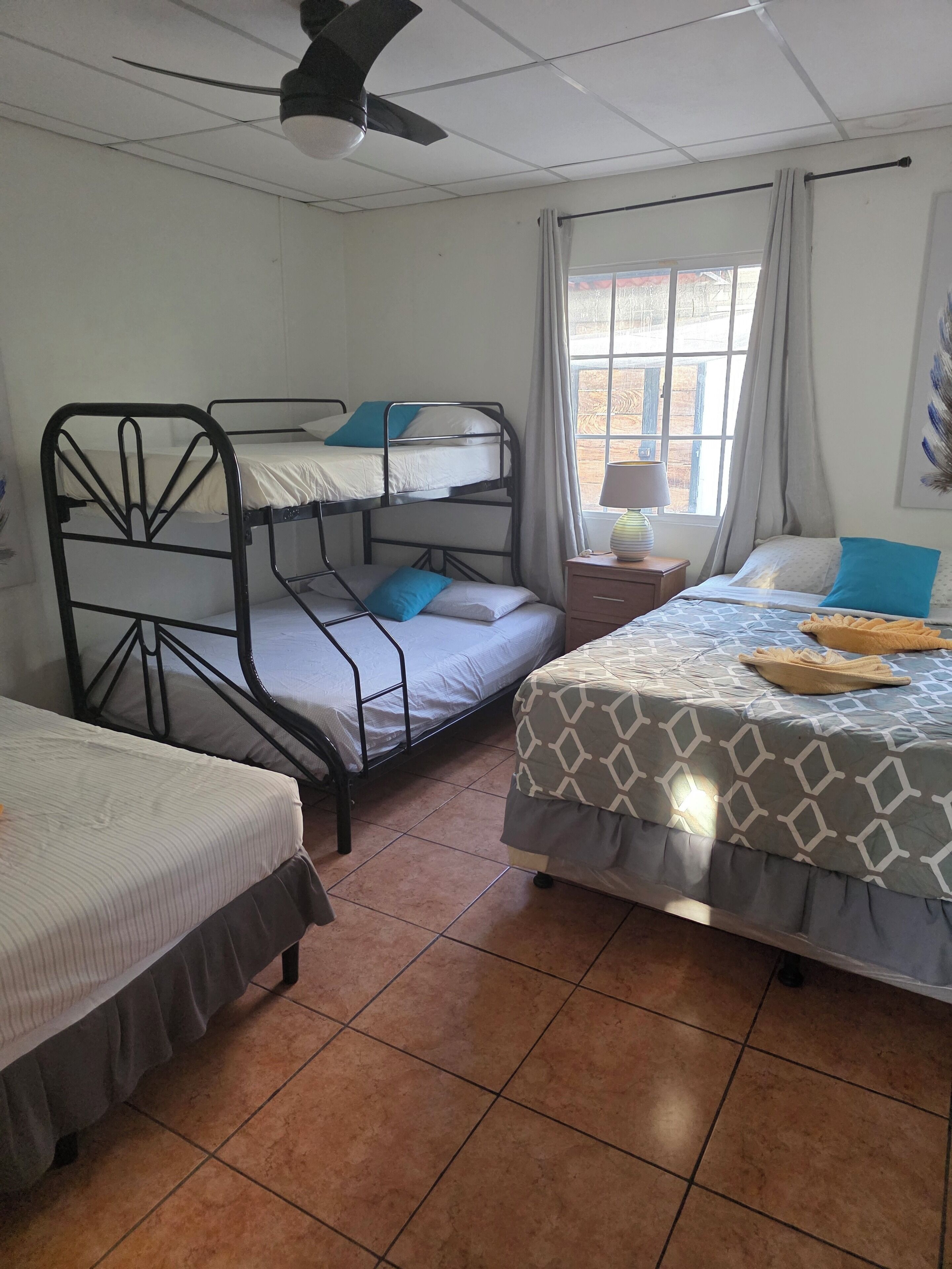 Un dormitorio con literas, ventilador de techo, ventana con cortinas y mesita de noche con lámpara.
