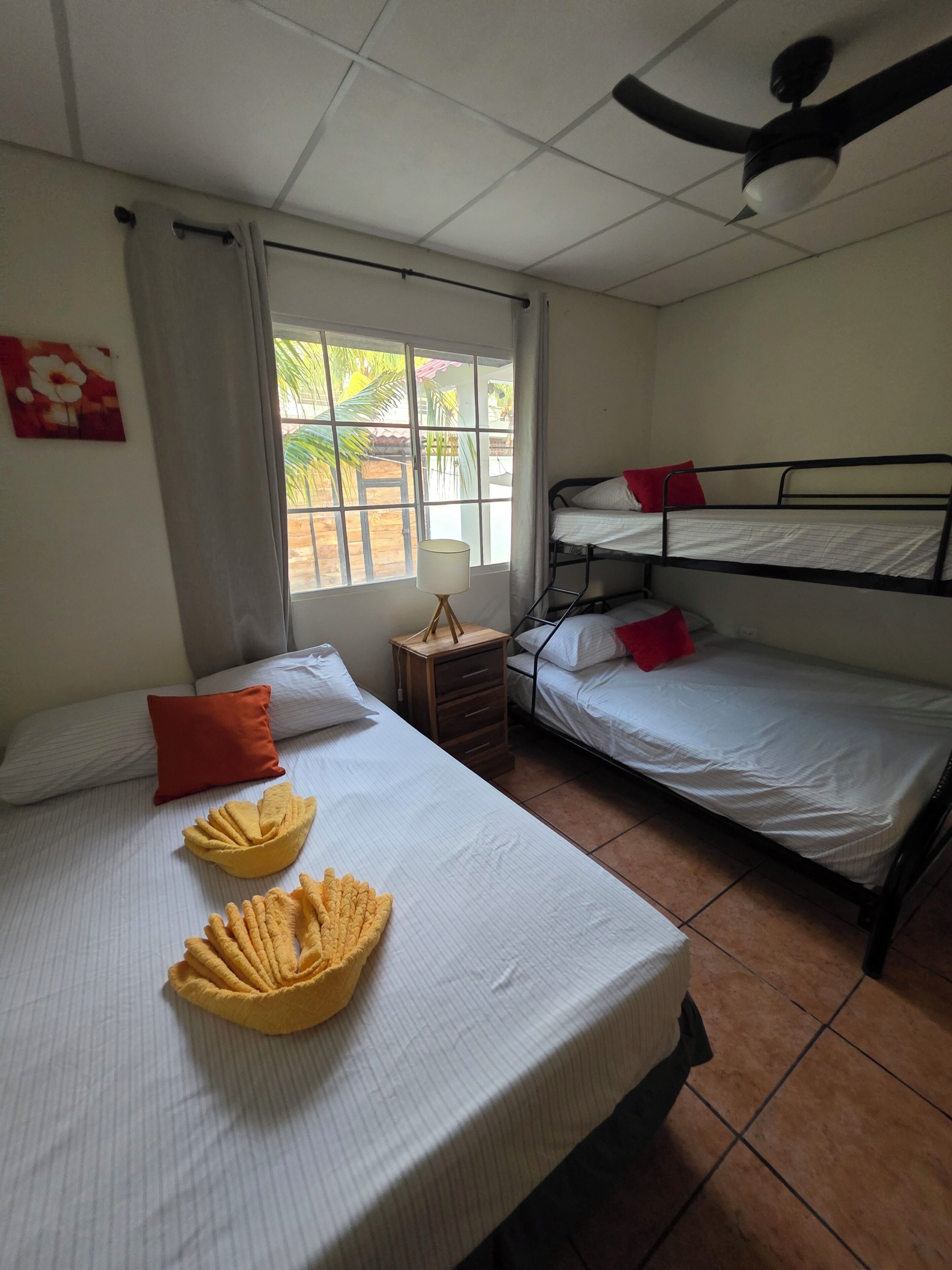 Una camera del dormitorio con due letti a castello, una finestra con tende, una ventola a soffitto e un tavolino con lampada.