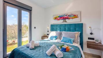 A bedroom with a large bed, a view through a sliding door, a painting on the wall, and a bedside table with a lamp.