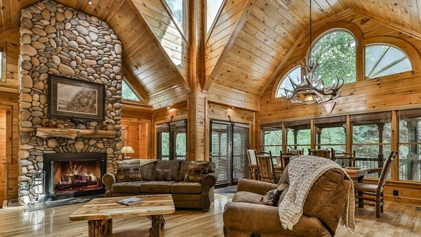 A log cabin interior with a stone fireplace, wooden furniture, and large windows. - Mountain Pooladise | Blue Ridge, Ga (Epworth)