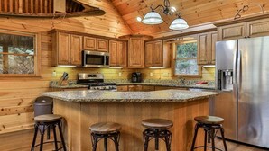 Ein Holzhütten-Interieur mit einer Küche, die eine Granitabdeckung, Barhocker, einen Edelstahlkühlschrank und eine Mikrowelle zeigt.