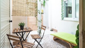 A small patio area with a wooden table and chairs, a small bed with a green mattress, and a window with a view of the outside. - Notti Magiche (Santa Maria di Castellabate)