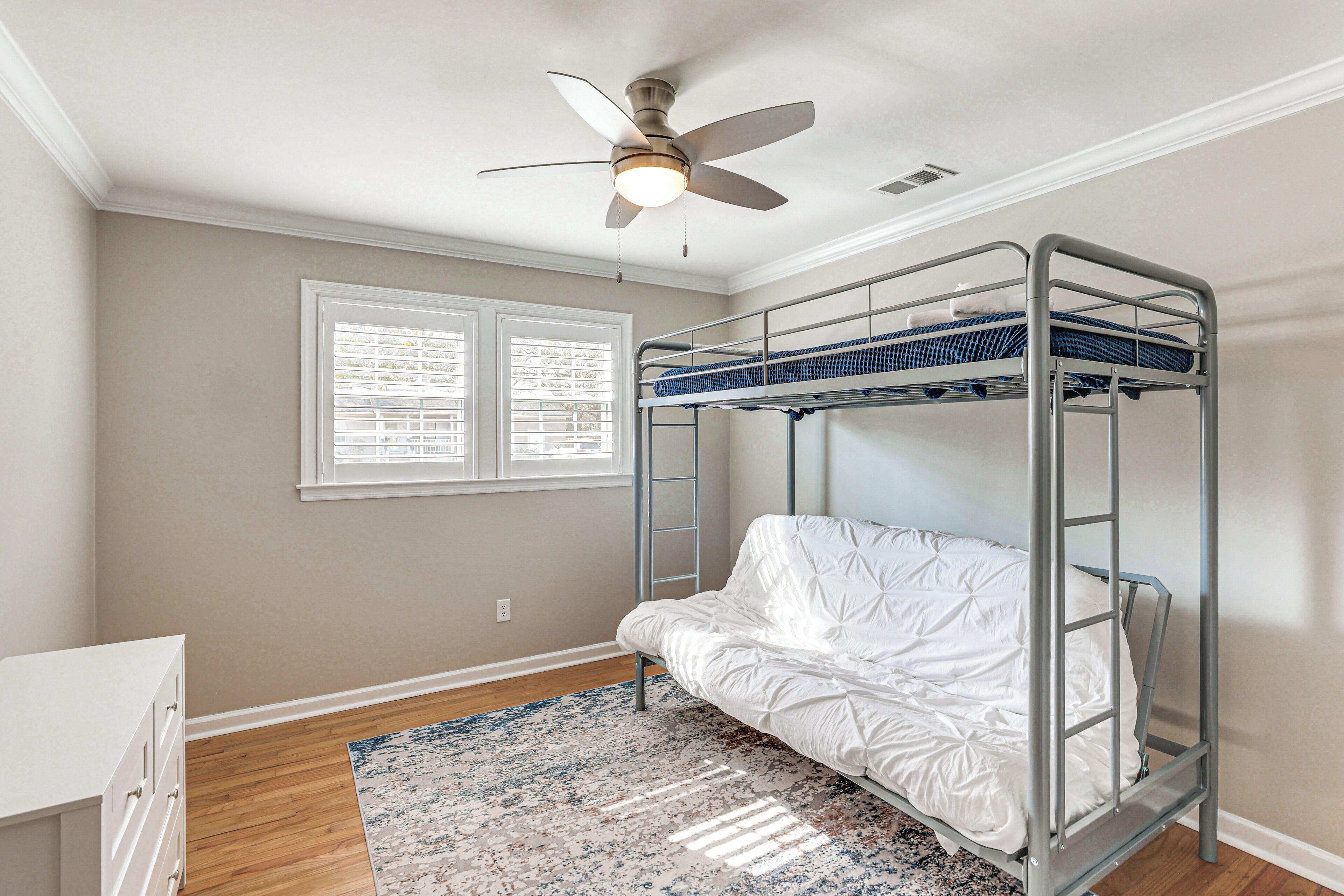 A bunk bed room with a ceiling fan, a window with blinds, and a white dresser.