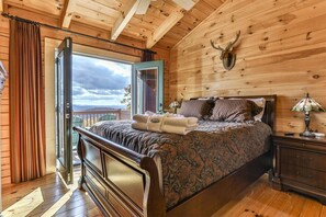 A wooden cabin bedroom with a bed, a nightstand, a lamp, a deer head decoration, and a view through a sliding door. - Skyview Retreat | Ellijay, GA (Ellijay)