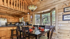 Interior de una cabaña de madera con comedor, cocina y ventanas amplias.