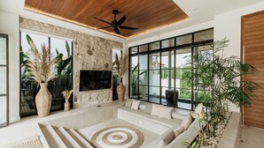 A modern living room with a stone wall, large windows, a ceiling fan, a flat-screen TV, and a seating area with a circular rug. - Luxurious 4 Bedroom villa with swimming pool in Uluwatu (Uluwatu)