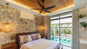 A bedroom with a stone wall, a ceiling fan, a bed with white bedding, a nightstand, and a view of an outdoor pool area. - Luxurious 4 Bedroom villa with swimming pool in Uluwatu (Uluwatu)