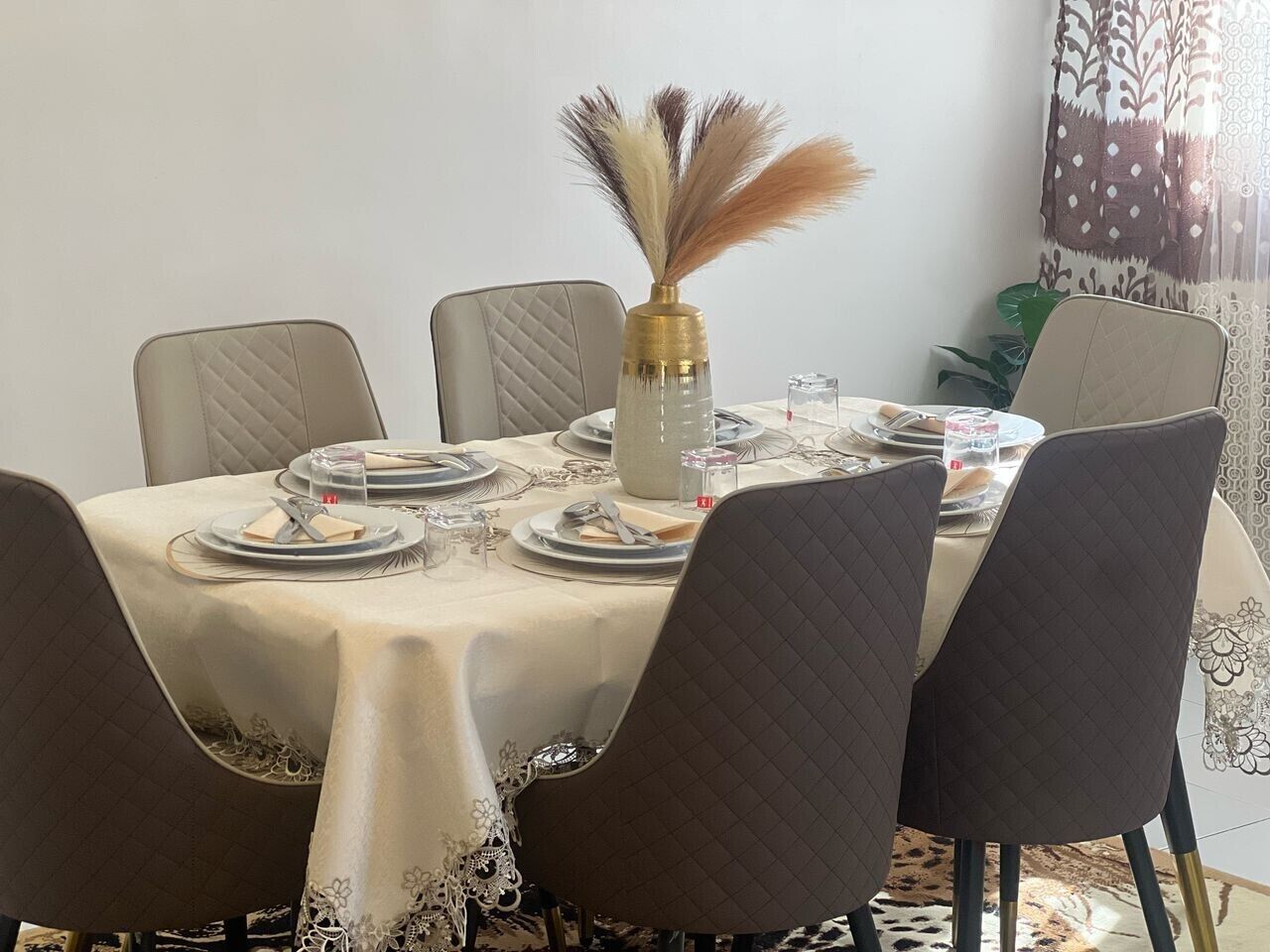 A dining table set for four with a vase of dried feathers, a patterned tablecloth, and a decorative wall piece.