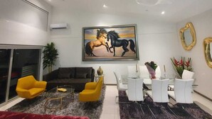 A modern living room with a large painting of horses, a sofa, yellow chairs, a coffee table, and a dining area with white chairs and a vase of flowers. - Penthouse safe and beautiful, with stunning views (San José)