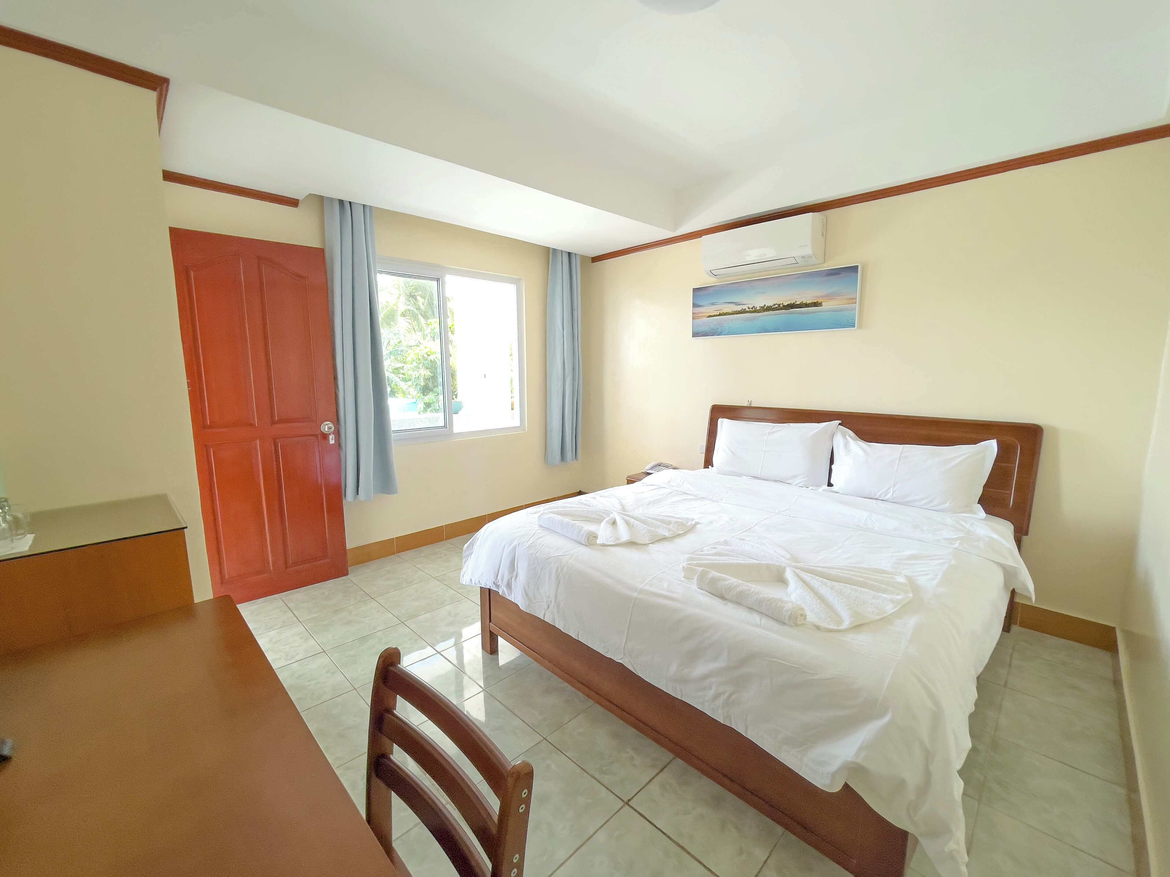 Habitación de hotel con una cama, una silla de madera, un escritorio, una ventana con cortinas y un cuadro en la pared.