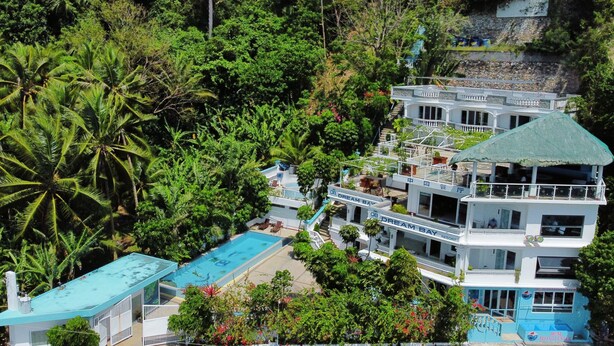 Un resort de varios pisos con piscina, rodeado de exuberante vegetación y palmeras.