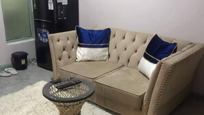 Un sofá seccional color beige con cojines azules y blancos, una mesa de centro de vidrio y un refrigerador negro al fondo.
