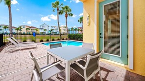 A poolside area with a table and chairs, a door with a window, and palm trees in the background.
