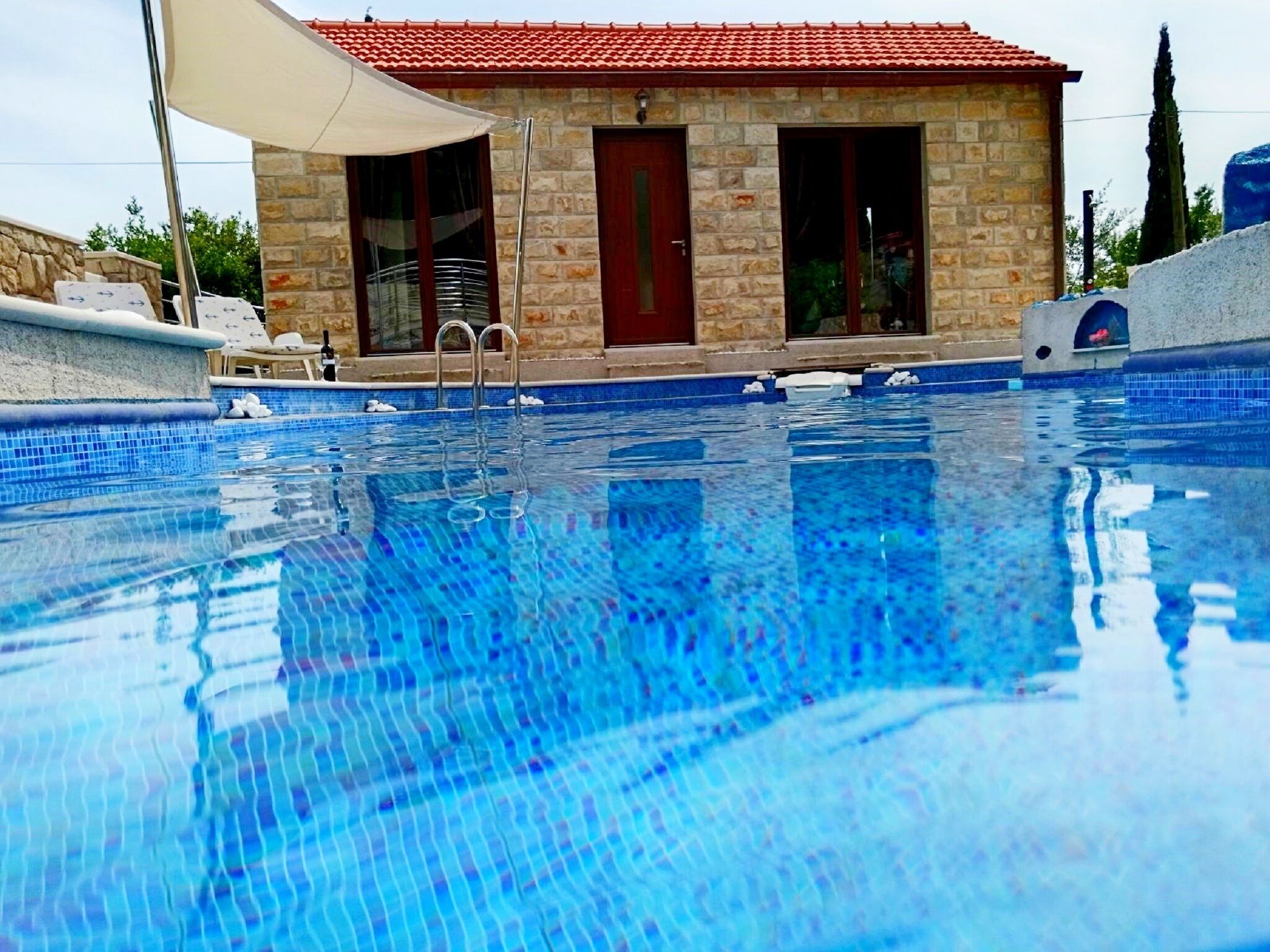 A swimming pool with clear blue water, a stone building with a red roof, and a white umbrella.