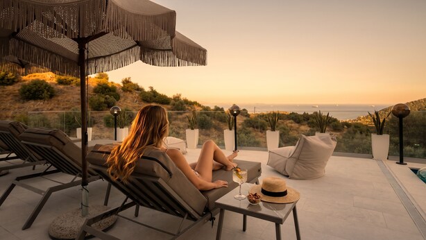A woman relaxing on a lounge chair with a view of the ocean and hills. - Saronic Citadel Luxury Villa | Private Estate on Salamina Island (Perani)