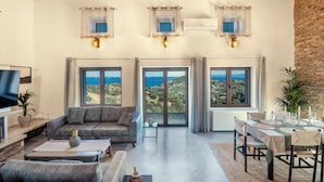 A spacious living area with a sofa, a dining table set for four, and large windows offering a view of the landscape. - Saronic Citadel Luxury Villa | Private Estate on Salamina Island (Perani)