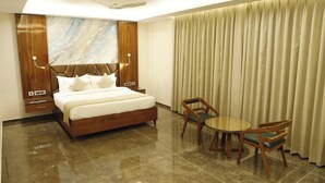 Una habitación de hotel con una cama grande, cabecero de madera, una obra de arte de mármol en la pared, un ventilador de techo, una mesa con sillas y un gran ventanal con cortinas.
