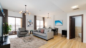 Un salon moderne avec un canapé gris, une table basse et un téléviseur. On y trouve également un coin repas avec une table et des chaises. La pièce est dotée de grandes fenêtres et offre une vue sur la ville.