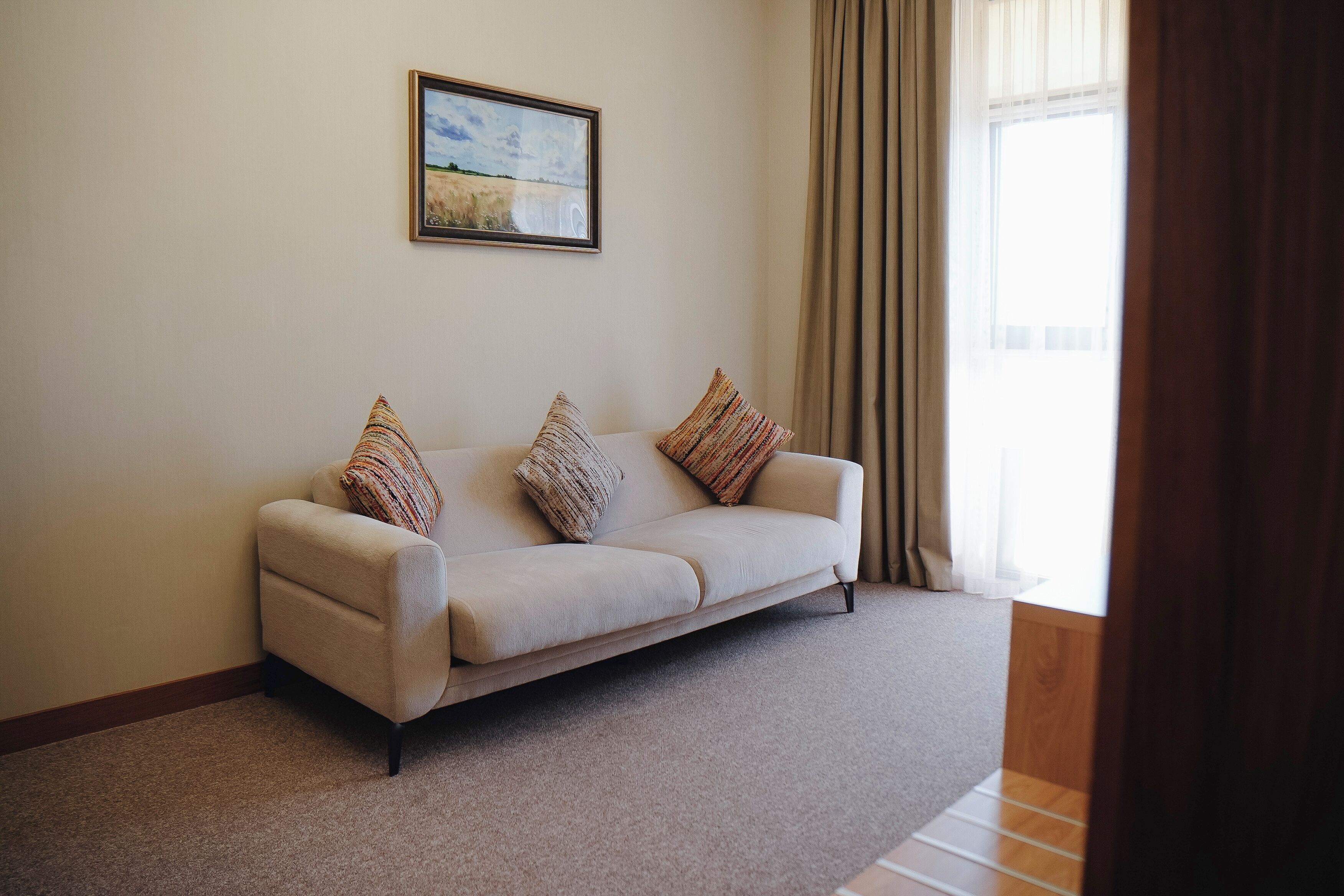 A living room with a beige sofa, patterned throw pillows, a framed landscape painting, and a large window with curtains.