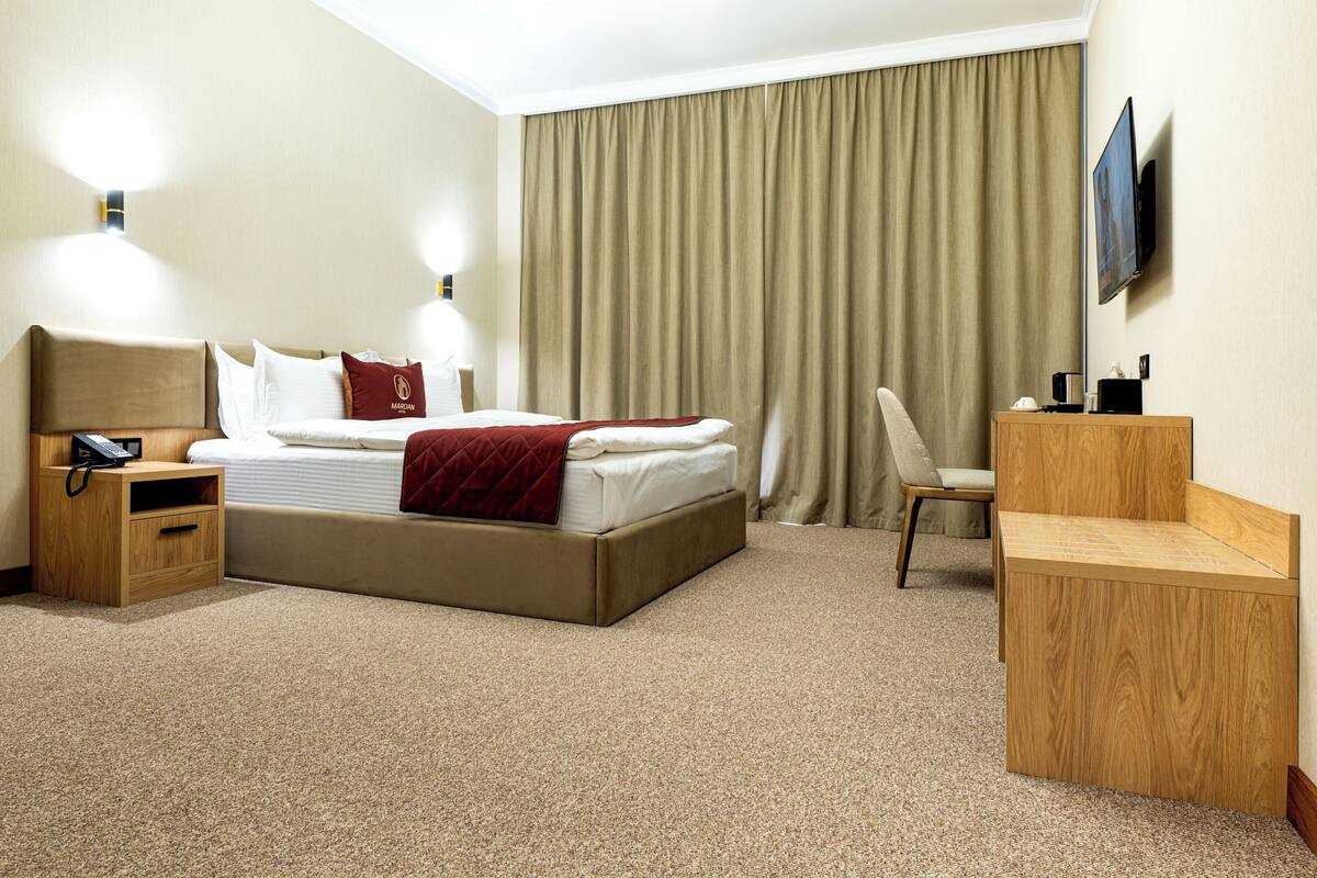 A hotel room with a bed, bedside table, lamp, TV, and a desk.