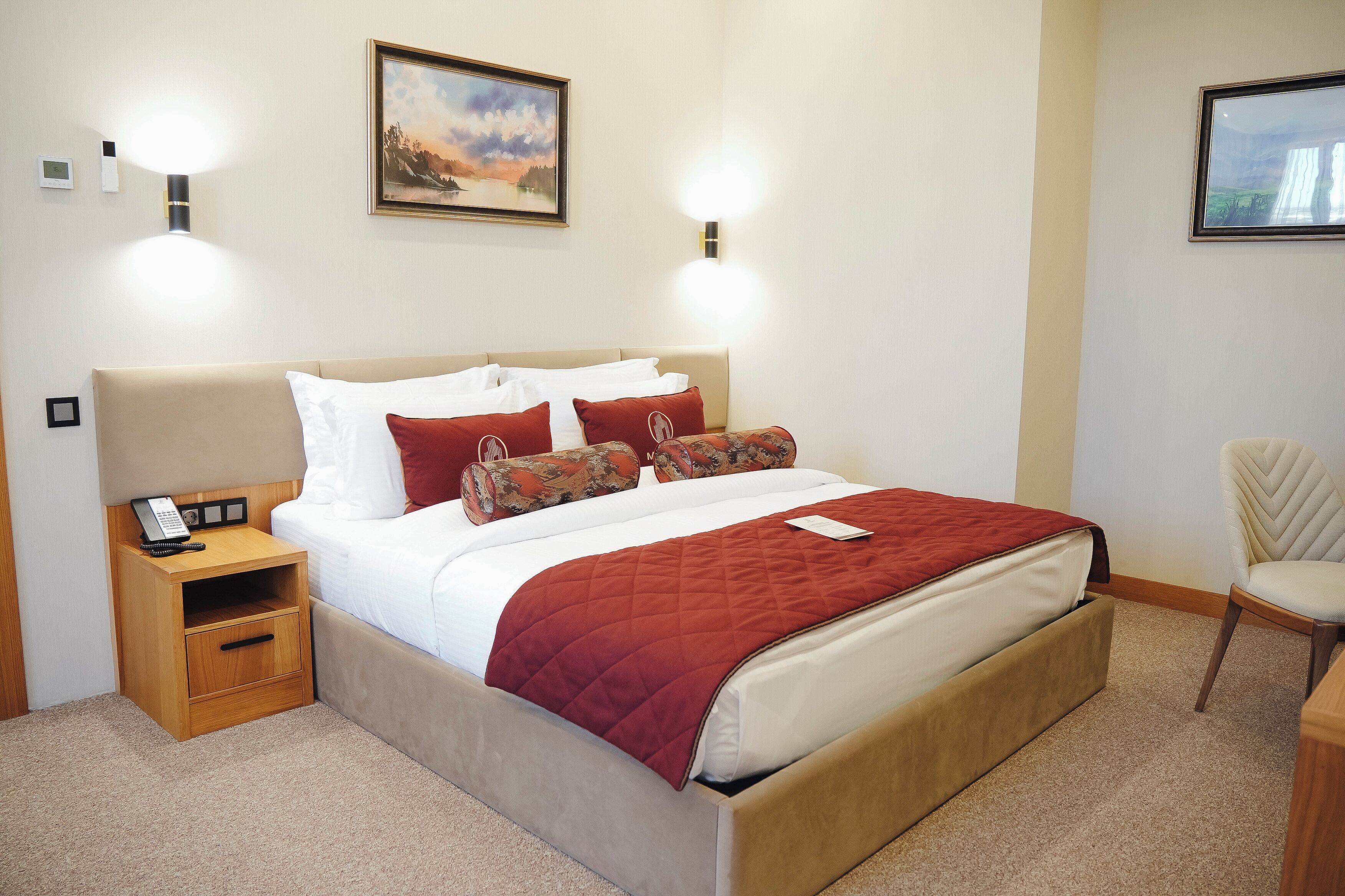 A neatly made bed with a red blanket, a bedside phone, and a small wooden nightstand.