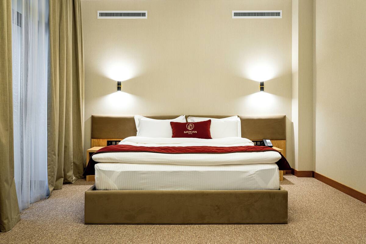 A hotel room with a large bed, two bedside lamps, a red pillow, and a wooden nightstand.