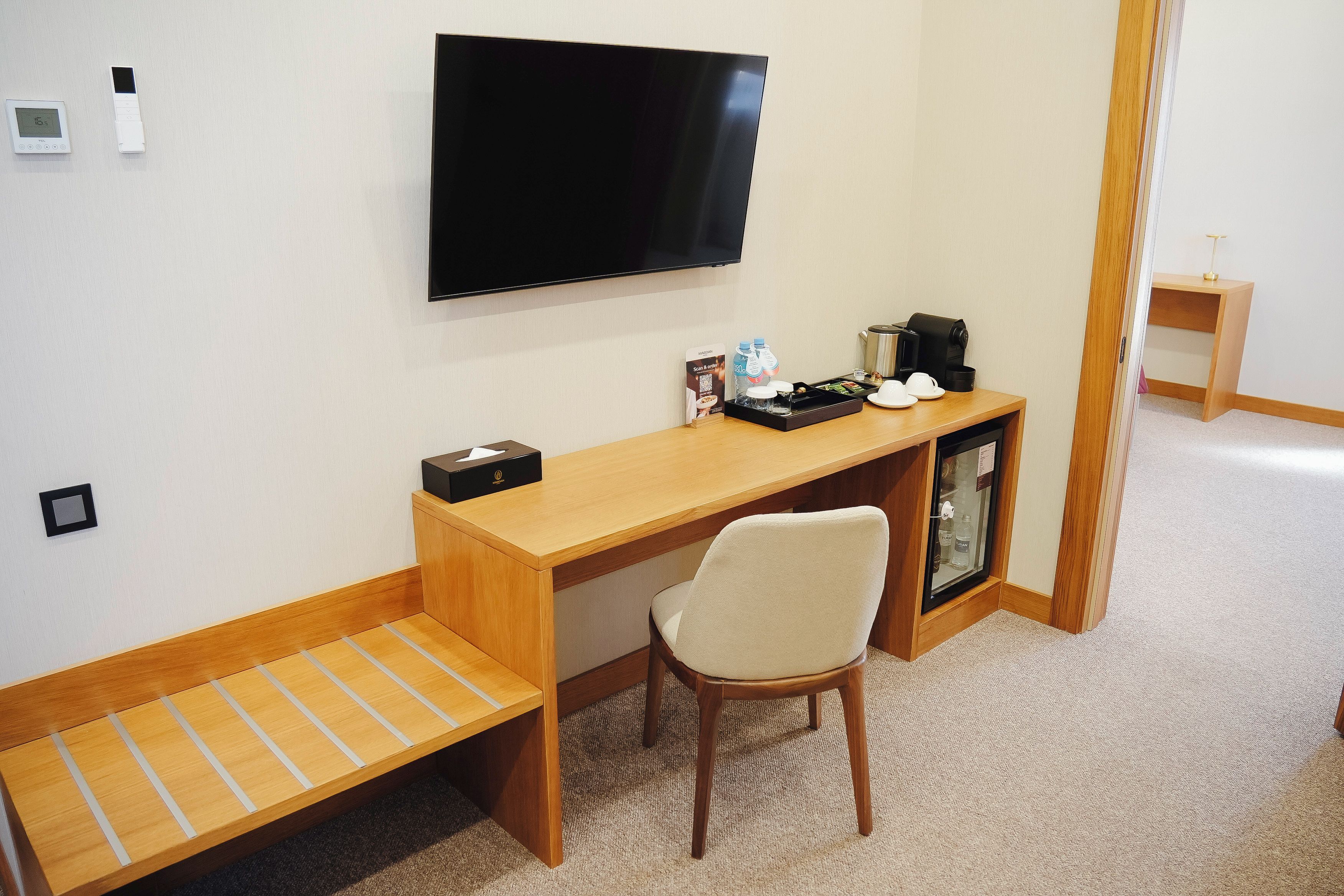 A hotel room with a wooden desk, a television mounted on the wall, a small cabinet with a coffee maker, and a chair.