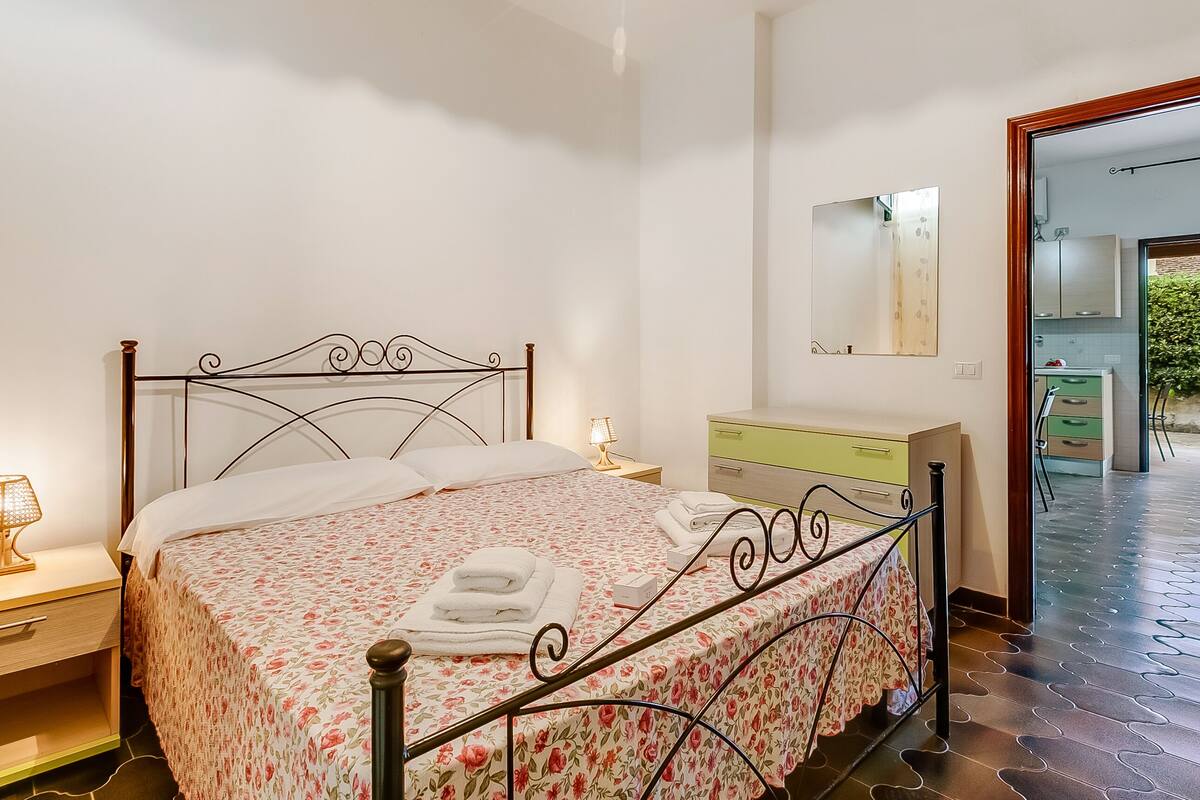 Un lit bien fait, recouvert d’une couvre-lit à motifs floraux, avec des serviettes soigneusement pliées, une lampe de chevet, une table de chevet et une commode avec un miroir.