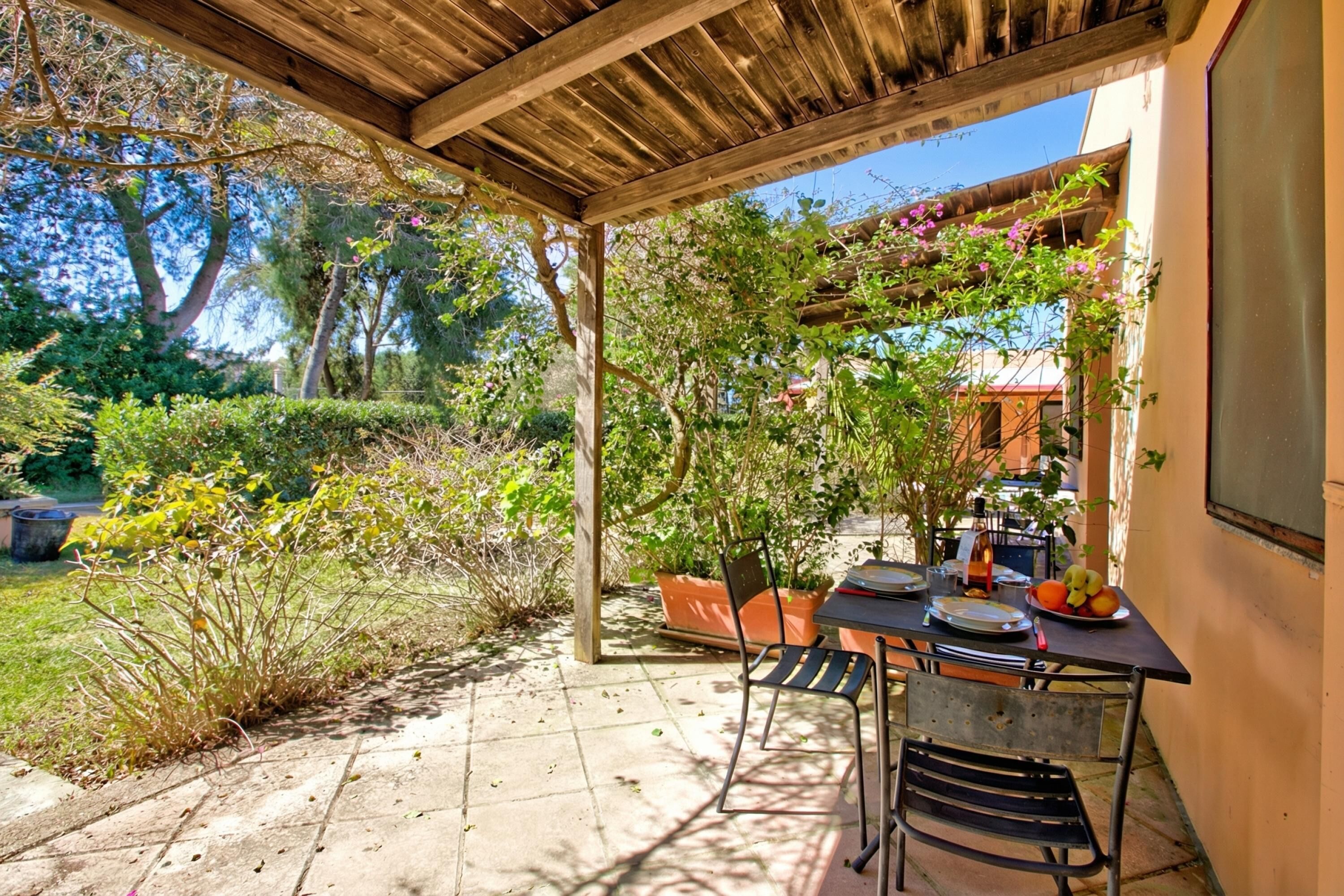 Eine überdachte Terrasse mit Esstisch, umgeben von Kübelpflanzen und Bäumen.