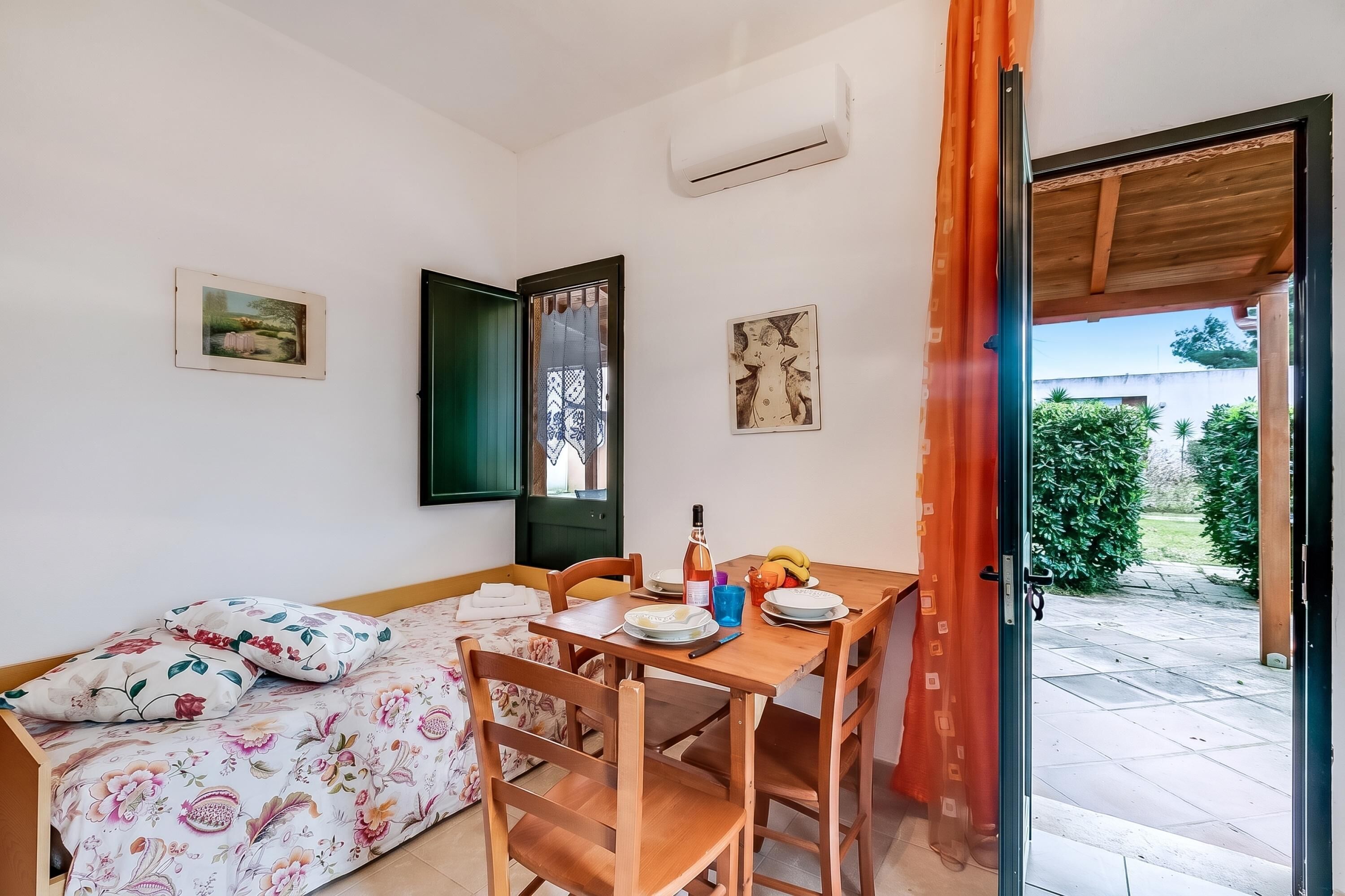 A room with a bed, a dining table with chairs, a window with a view of greenery, and a door leading outside.