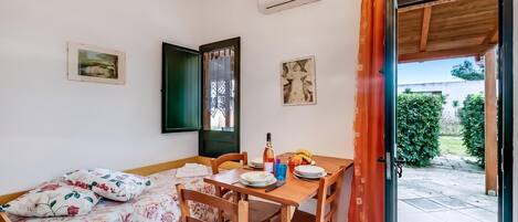 A room with a bed, a dining table with chairs, a window with a view of greenery, and a door leading outside.