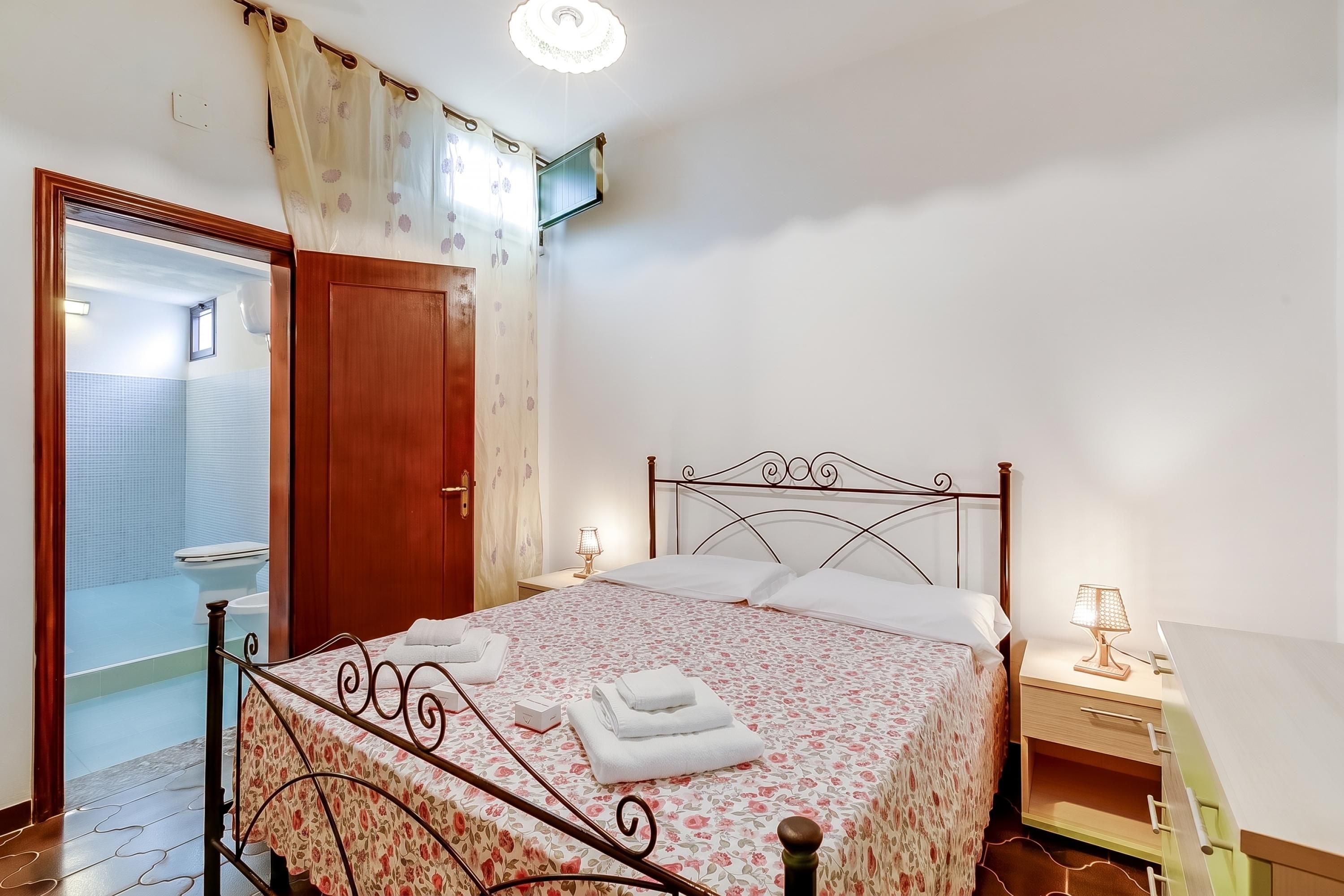 Une chambre à coucher avec un lit, une table de chevet, une lampe et une salle de bain visible à travers une porte ouverte.