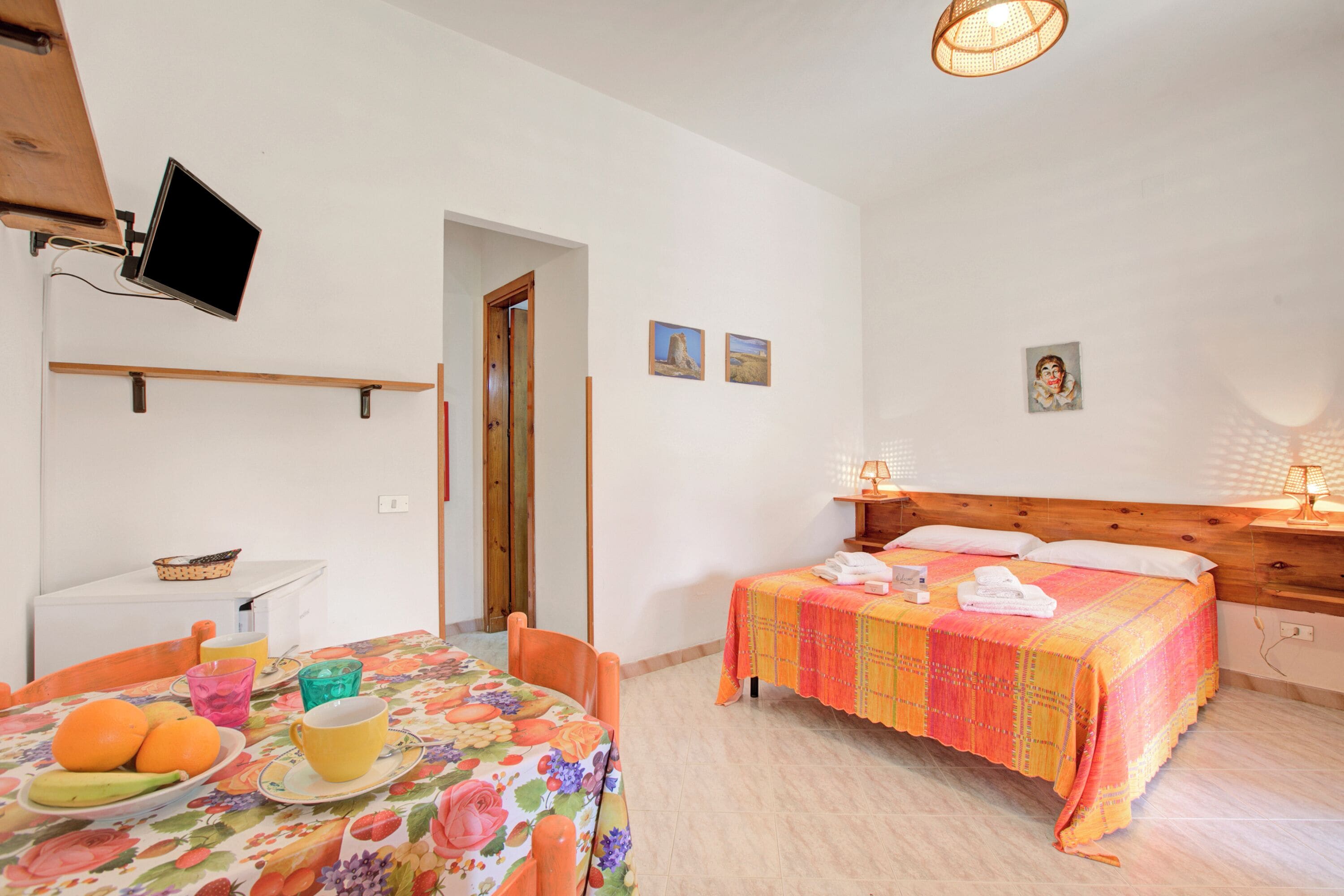 Um quarto com uma cama, uma mesa com frutas, uma televisão e uma porta.
