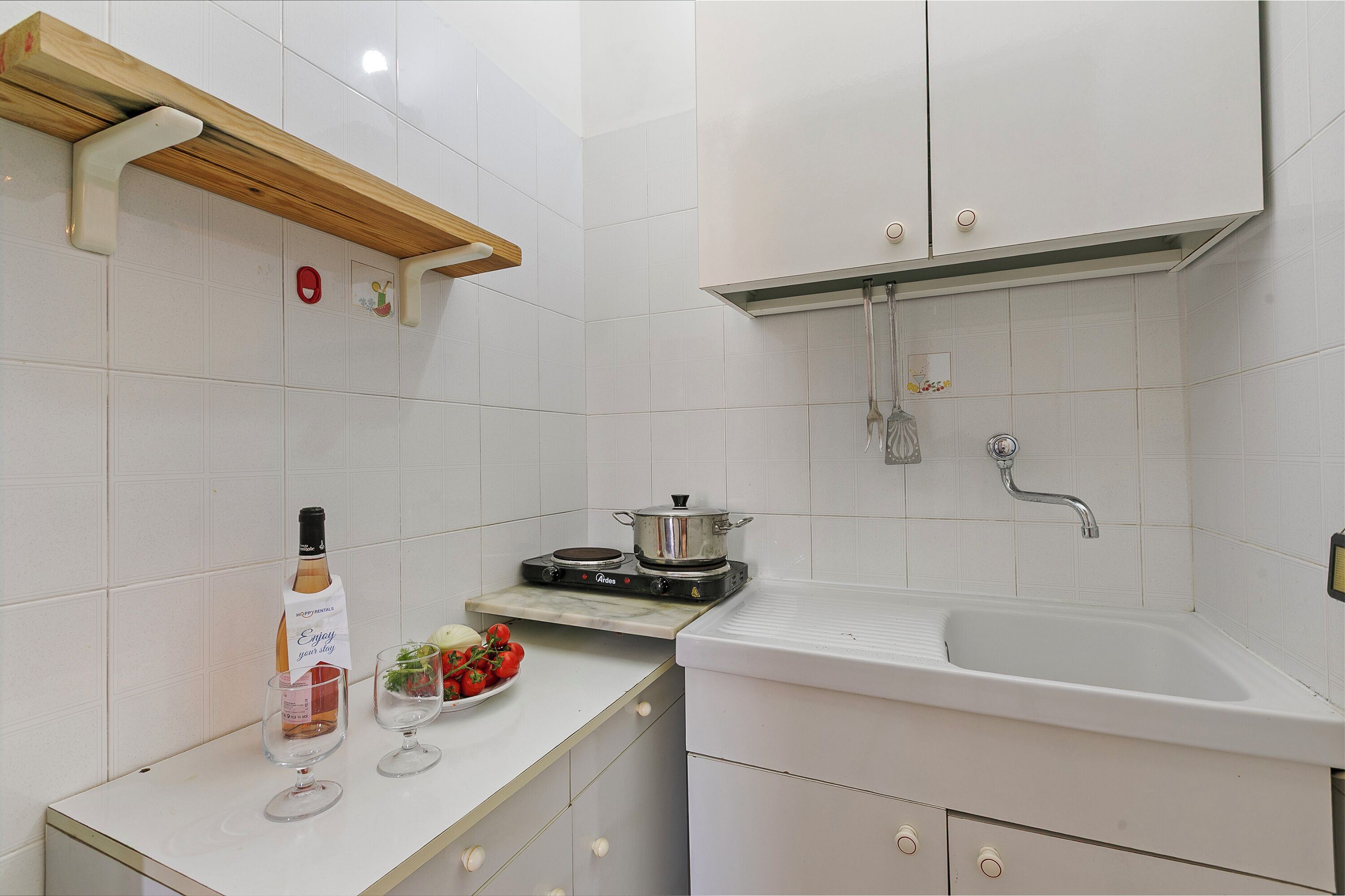 Une cuisine avec des meubles blancs, une étagère en bois, une cuisinière, un évier et une bouteille de vin.