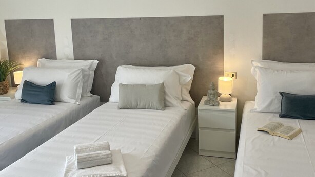 Quarto de hotel com três camas de solteiro, cada uma com roupa de cama e almofadas brancas, uma pequena mesa de cabeceira com uma luminária, e uma estátua sobre uma das mesas.