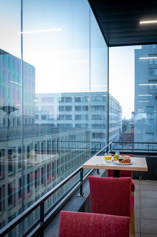 A modern dining area with a table set for two, a glass railing, and a city view through large windows.