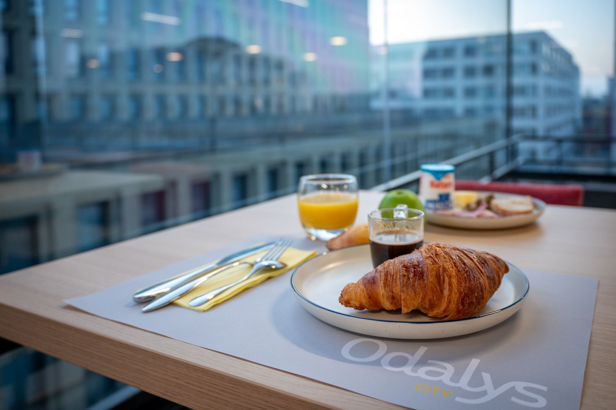A breakfast setup with a croissant, juice, and a plate of food on a table with a view of a cityscape.