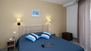 Une chambre d’hôtel avec un lit, deux lampes de chevet, un ordinateur portable, une petite table avec une tasse et des verres, et un tableau accroché au mur.