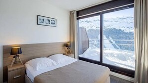 A hotel room with a bed, two bedside lamps, a framed picture on the wall, and a large window with a snowy mountain view.