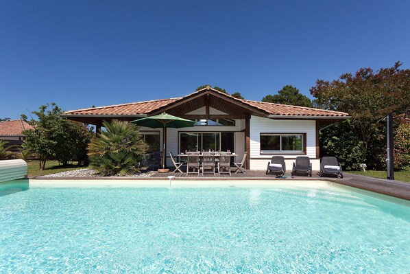 A house with a swimming pool, a patio with chairs, and a covered area with a table and chairs.