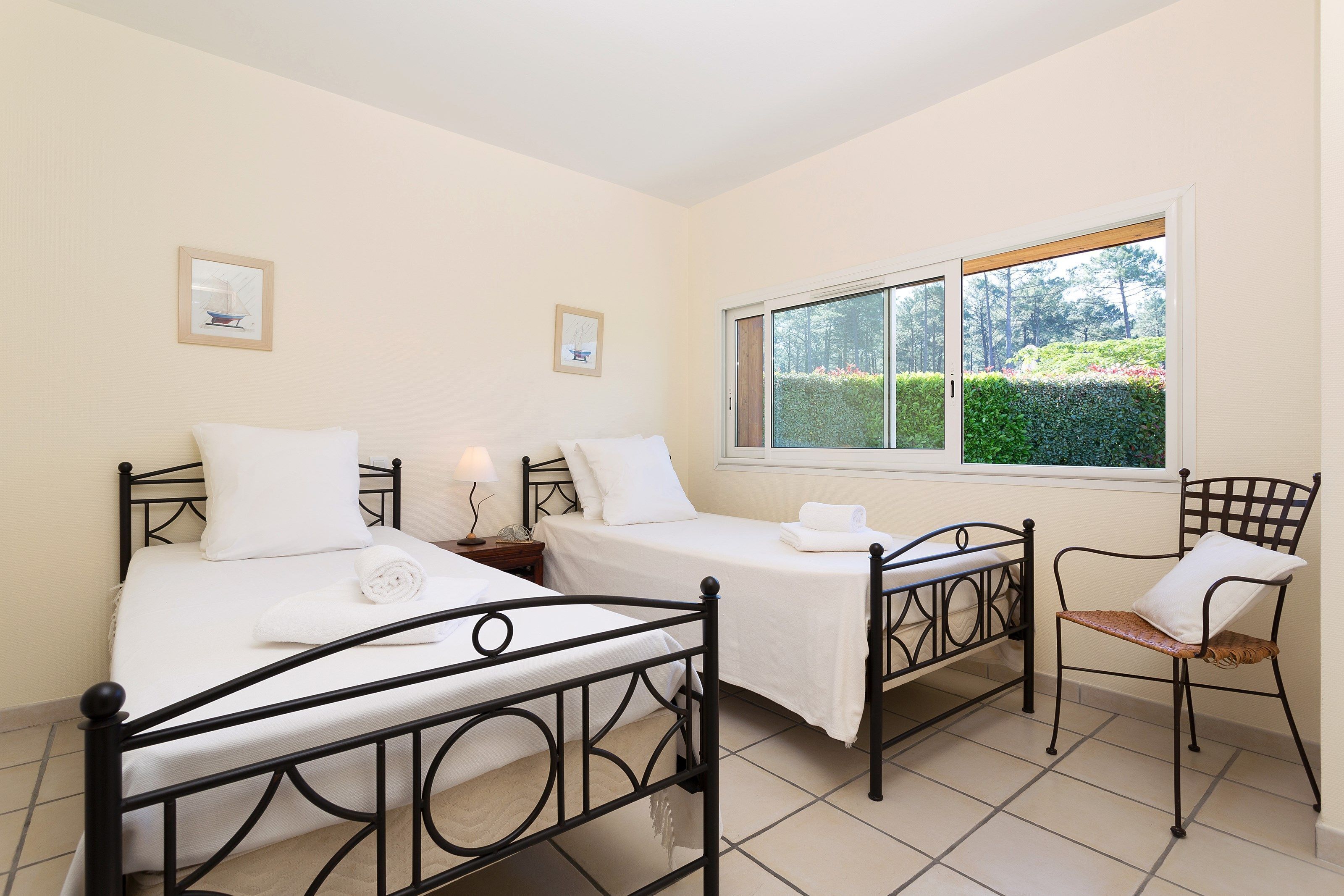 A bedroom with two beds, a chair, and a window with a view of greenery.