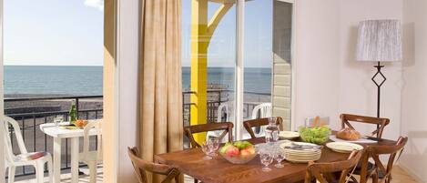 A dining area with a view of the sea, a table set with plates, glasses, and a bowl of fruit.