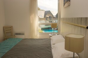 Une chambre d’hôtel comprenant un lit, une lampe de chevet, une chaise, une fenêtre donnant sur une piscine et des bâtiments, ainsi qu’une lampe fixée au mur.