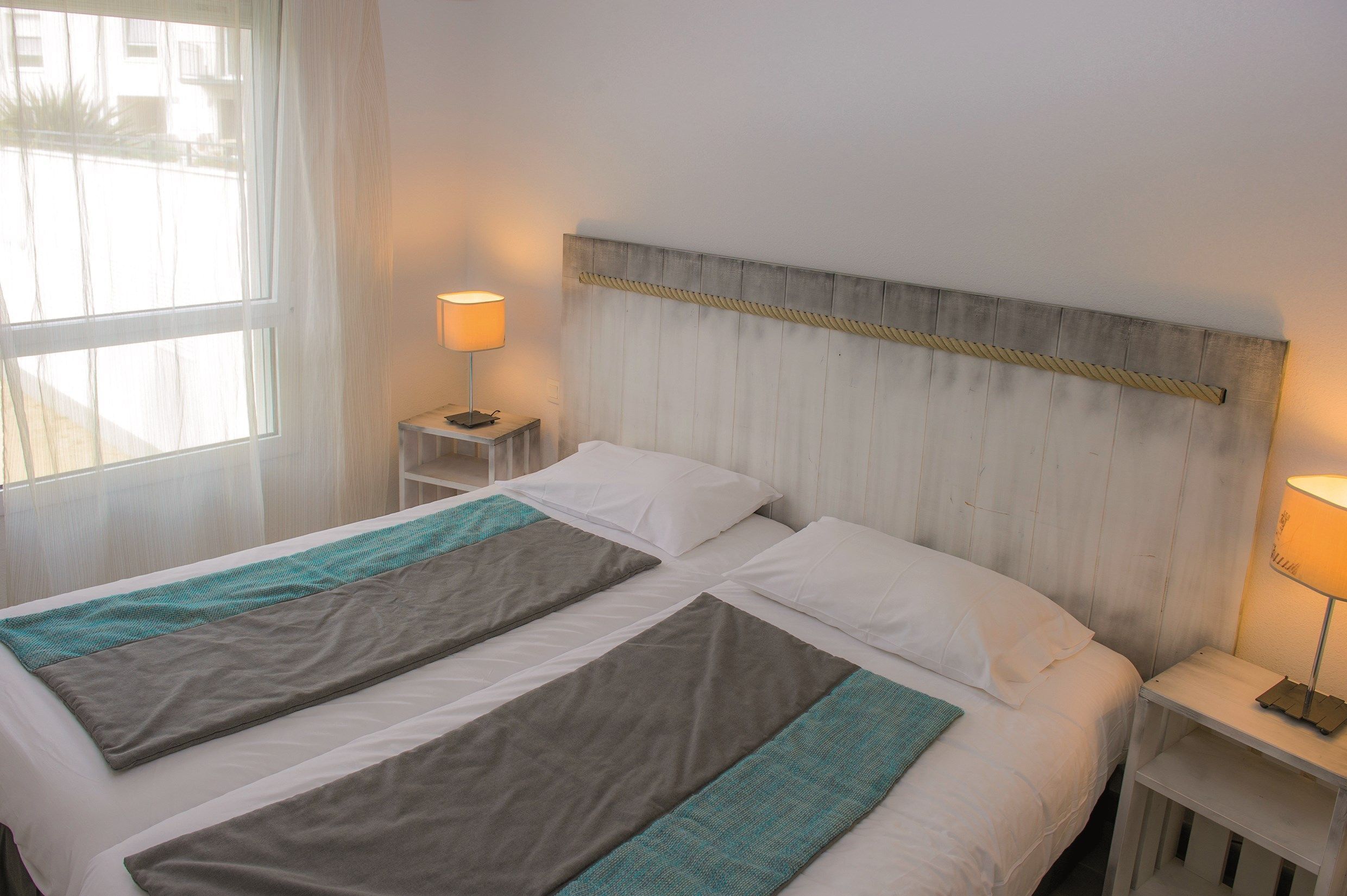 A neatly made bed with a headboard, two bedside tables with lamps, and a window with sheer curtains.