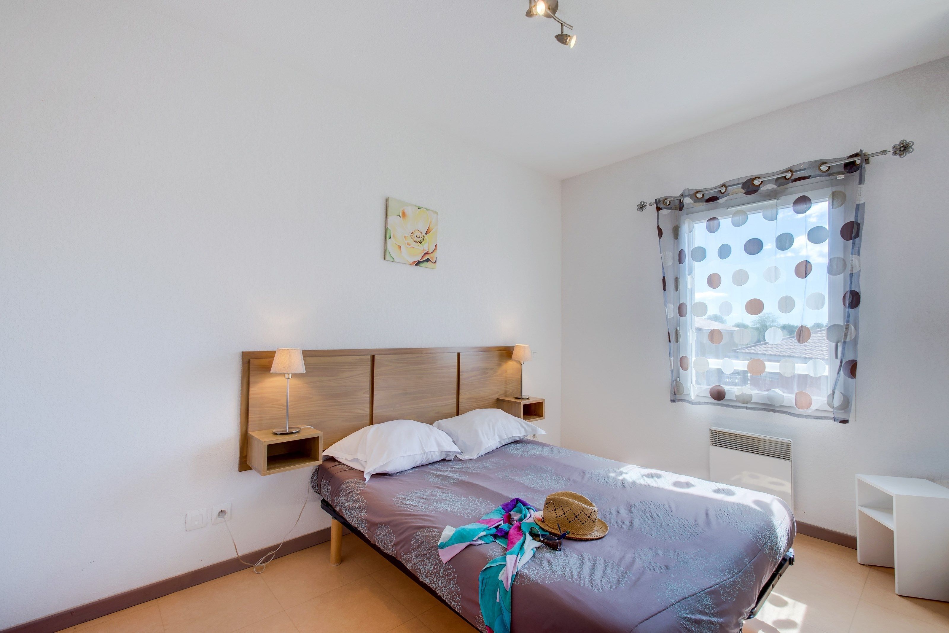 A bedroom with a bed, bedside lamps, a window with a polka dot curtain, a wall-mounted artwork, and a small white table.
