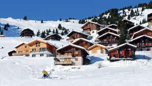 A cluster of wooden chalets on a snowy mountain slope, with ski tracks and a snowmobile visible.