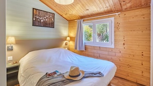A cozy wooden room with a bed, two wall-mounted lamps, a window with curtains, and a framed picture on the wall.
